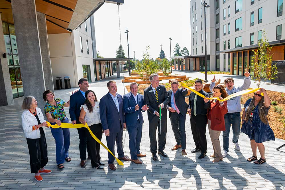 A group of people cutting a ribbon in front of a newly opened building with students and dignitaries