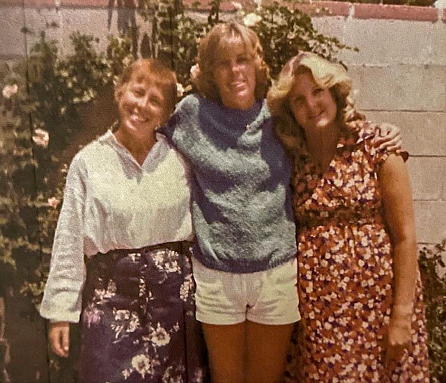 Three women standing side by side in front of a textured brick wall, smiling and engaging with each other