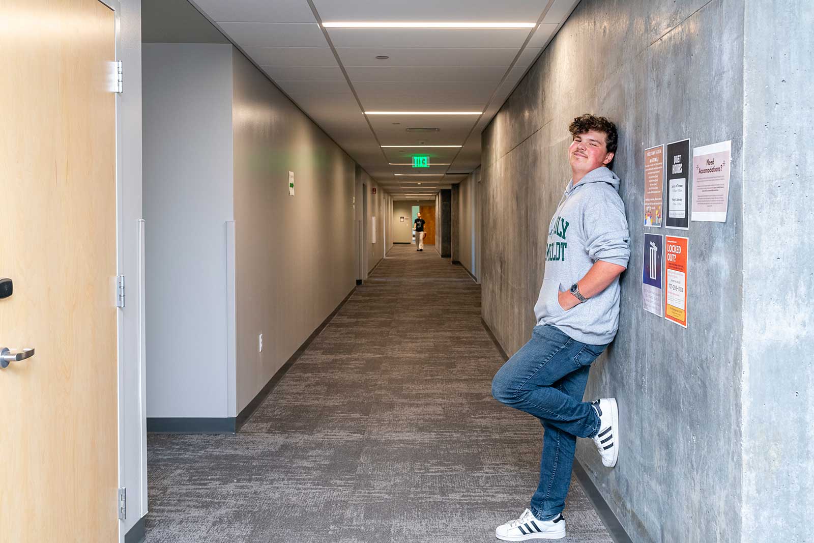 A student leans against a wall in a hallway, appearing relaxed and contemplative