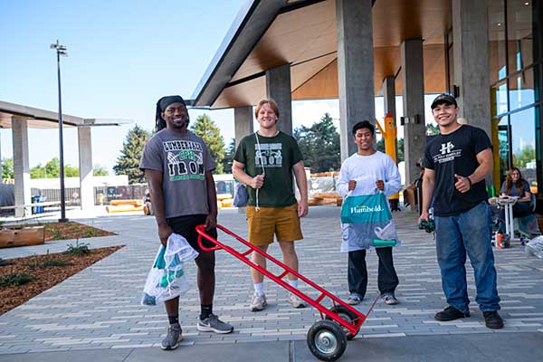 Four people stand in front of a building beside a hand truck, engaged in conversation and looking towards the camera