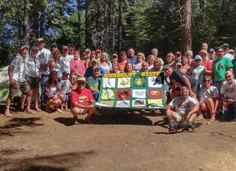  Jackson Meadows, located north of Lake Tahoe, is the backdrop for a memorable photo of the group. They are holding a banner created by Cathy Flores, showcasing a variety of t-shirts designed for their trips. Flores’ favorite shirt is a bright red one featuring an illustration of a campfire and people gazing at the stars.